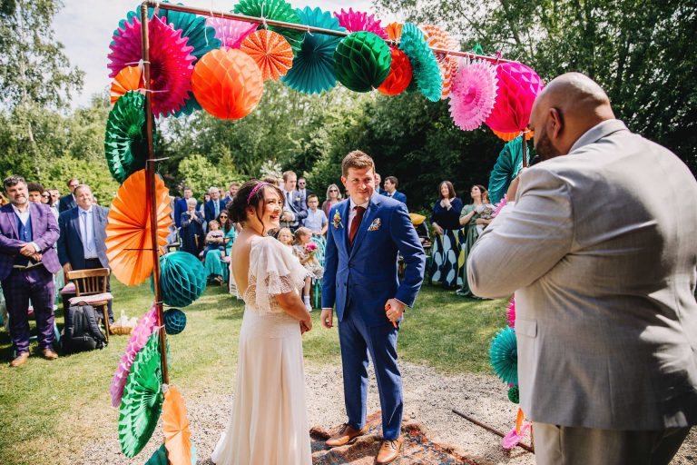 couple standing at the front of their ceremony in the sunshine at Acorn Barn. Surrounded by a flower arch